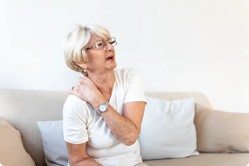 Senior woman experiencing menopause joint pain while holding her sore shoulder on a couch.