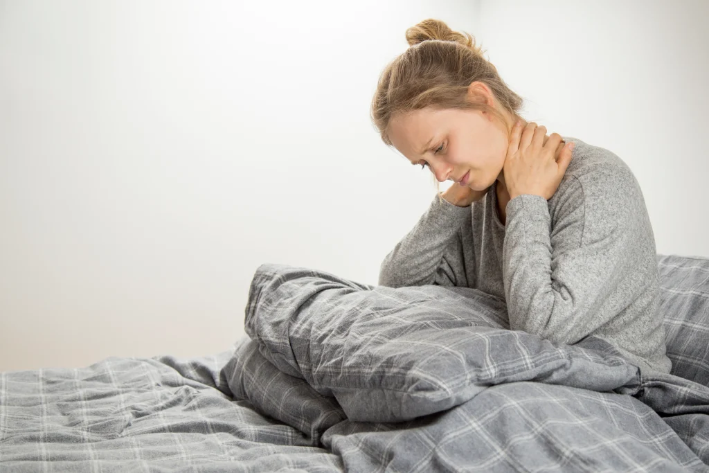 A woman sitting in bed rubbing her sore neck dealing with joint and muscle pain at night.