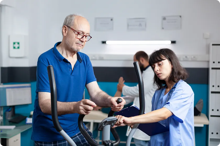 Elderly man exercising on a stationary bike with nurse monitoring therapy for dementia patients.