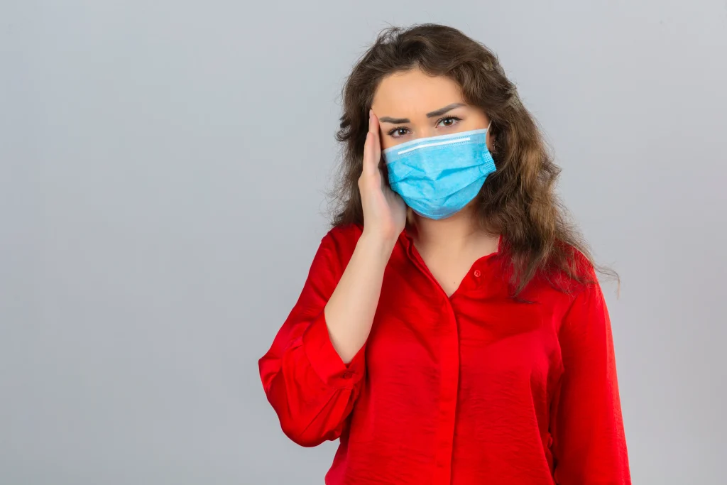 Woman wearing a surgical mask holds her forehead identifying signs of nasal polyps.